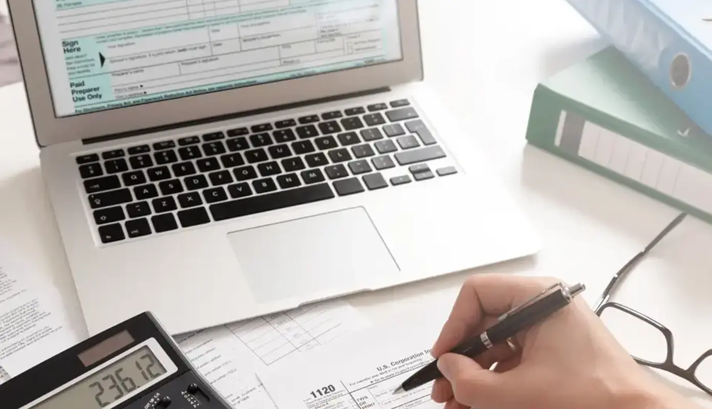 Close-up of hands filling out a tax form on a laptop, with a calculator displaying '236.12' and other tax documents nearby.